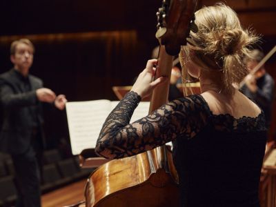 Student playing the double bass on stage with a conductor in the background